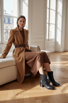 Canna black leather Chelsea ankle boots styled with a belted camel wool coat and chocolate knit dress in a sunlit classic interior