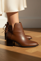 Close-up detail of Attalea brown leather ankle boots showing the hand-tied leather bow, ankle strap stitching and black stacked heel