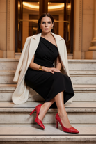 Callisia red studded leather pumps styled with a black asymmetric draped column dress and ivory wool coat on marble steps at dusk